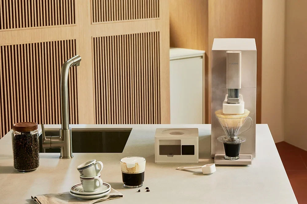 STEAM 100 Barista 净水器摆在极简风格类厨房空间的浅色台面上。可见一台带玻璃锥形滤杯的白色透明高身咖啡机、一个白色小盒、一罐咖啡豆、两只叠放的陶瓷杯,以及一只盛有深色咖啡的小玻璃杯。