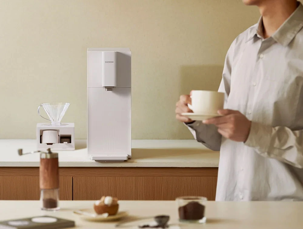 身穿浅灰色衬衫的人在台面上端着一只白色杯子,旁边是 STEAM 100 Barista 净水器和小型手冲滤杯,前景有咖啡豆和磨豆机。